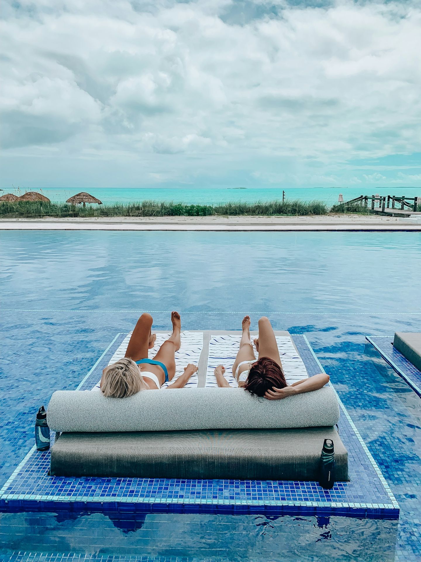2 women lying on blue towel on blue swimming pool during daytime in front of the beach